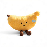 Peluche banana gialla dal viso sorridente, con zampe marroni, posato su sfondo bianco.
