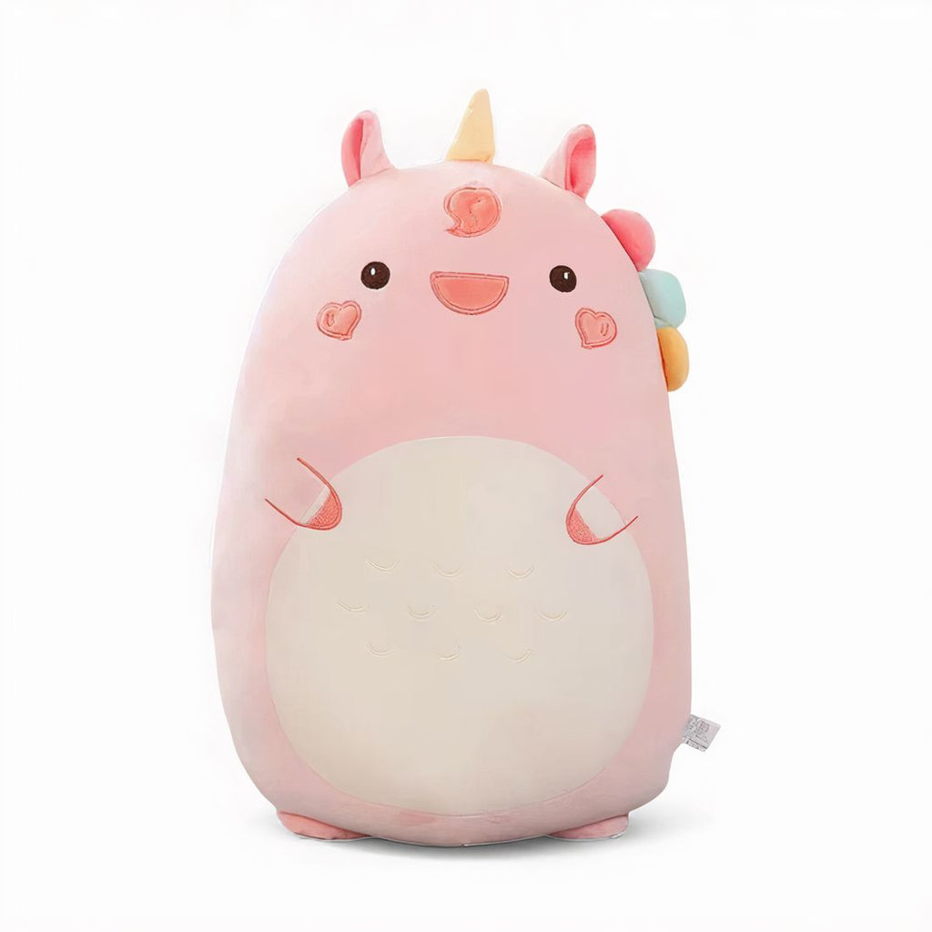 Grande cuscino peluche unicorno rosa, con corno giallo, criniera multicolore, guance a forma di cuore e grande pancia color crema, appoggiato su un letto in una cameretta luminosa.