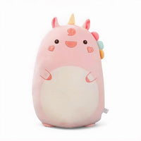 Grande cuscino peluche unicorno rosa, con corno giallo, criniera multicolore, guance a forma di cuore e grande pancia color crema, appoggiato su un letto in una cameretta luminosa.